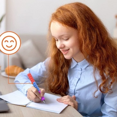 Criança ruiva a escrever num caderno aberto numa secretária com computador e planta.