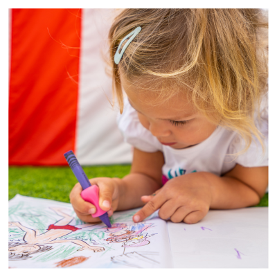 Criança loira a colorir um desenho, relva verde e tecido vermelho e branco ao fundo