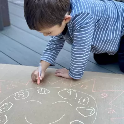Criança a desenhar com giz colorido em cartão castanho no chão de madeira