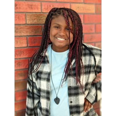 Pessoa sorridente com tranças vermelhas e pretas, casaco xadrez e colar preto em forma de coração junto a uma parede de tijolos.