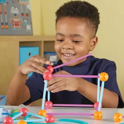 Criança a montar brinquedo de construção com bastões roxos, hastes azuis e esferas vermelhas e laranja numa mesa branca.