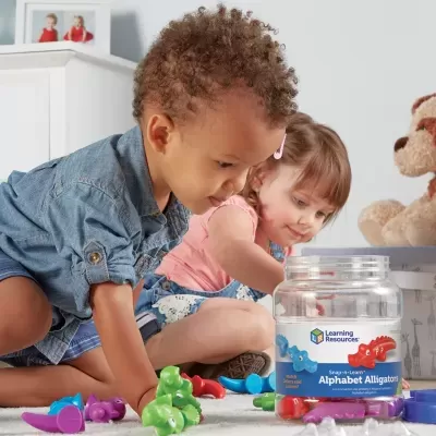 Frasco transparente do produto Alphabet Alligators com peças plásticas coloridas e crianças a brincar no chão