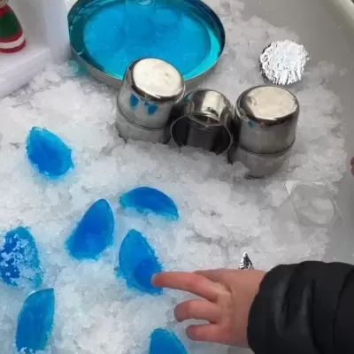 Recipiente branco com gelo, pedaços azuis translúcidos, copos metálicos prateados, prato azul e mão infantil