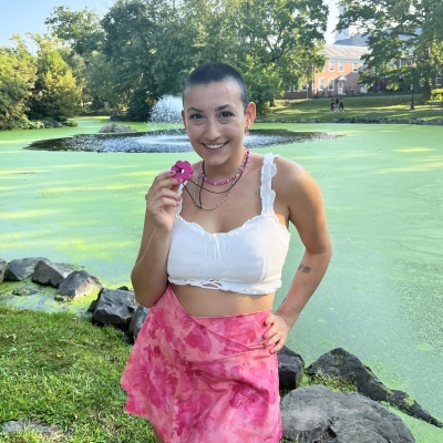 Mulher vestindo top branco e saia rosa com padrão tie-dye junto a lago verde e pedras