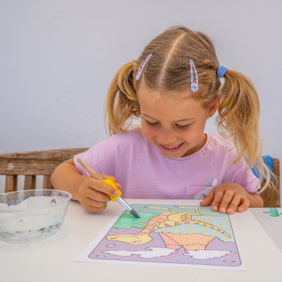 Criança a pintar um desenho de dinossauro numa mesa branca com tigela de água ao lado