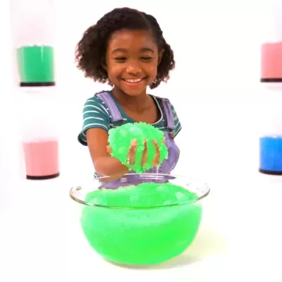 Criança a brincar com slime verde num recipiente de vidro, com frascos coloridos na parede ao fundo.