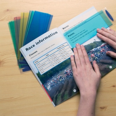 Livro aberto com informações sobre corridas e réguas coloridas transparentes ao lado em mesa de madeira.