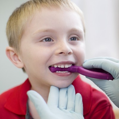 Criança loira com camisa vermelha a ser examinada com espelho bucal roxo por médico com luvas verdes