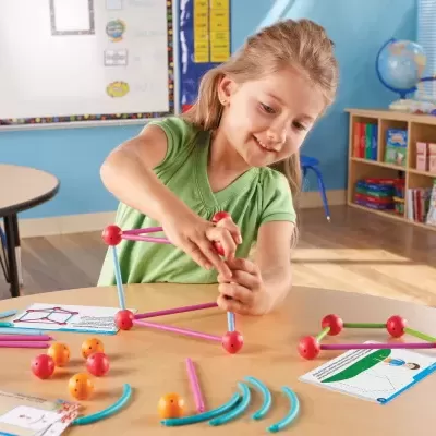 Criança a montar figuras geométricas com palitos e bolas coloridas numa sala de aula