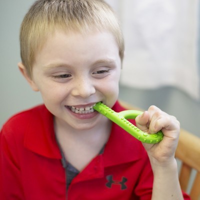 Criança loira a morder brinquedo verde com textura bolha, vestindo polo vermelha.