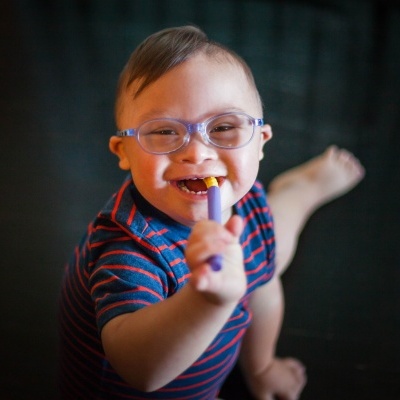 Criança sorridente com óculos azuis e camisa de riscas segurando objeto roxo e amarelo