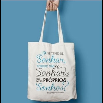 Saco de pano branco com texto impresso colorido, segurado por uma mão contra fundo azul