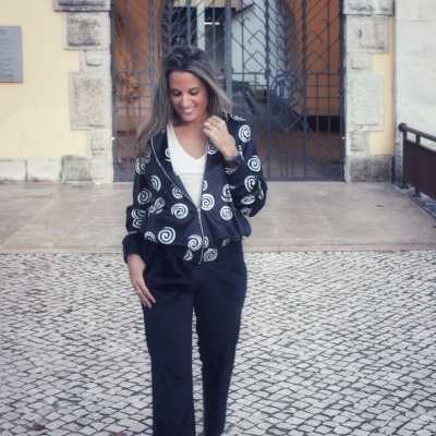Mulher com casaco preto de espirais brancas, calças pretas largas e sapatos pretos em calçada de pedra.