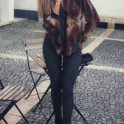 Mulher com calças pretas justas e camisa transparente com padrão abstrato em tons castanho e preto num espaço exterior com cadeiras de esplanada.