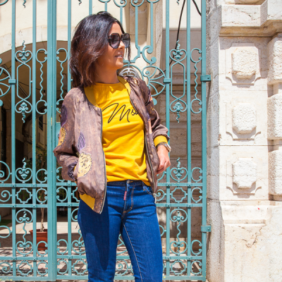 Mulher com casaco bomber castanho e t-shirt amarela à frente de portão metálico azul