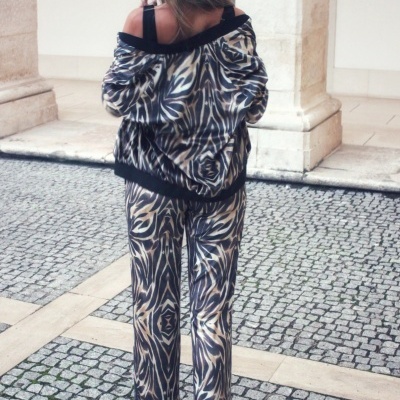 Pessoa de costas com conjunto de roupa padrão abstracto castanho, branco e preto, casaco e calças, usando ténis brancos coloridos em piso de calçada portuguesa.