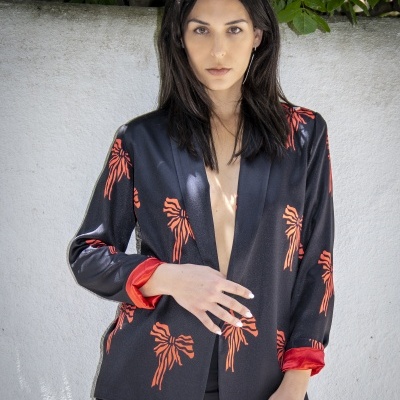 mulher com blazer preto de laços cor de laranja e forro vermelho junto a parede branca