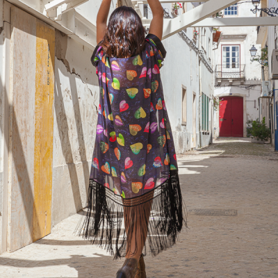 Mulher de costas a usar uma roupa exterior preta colorida com franjas, numa rua citadina.