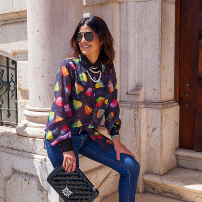 Mulher sentada com camisa estampada de gotas coloridas, calças de ganga azuis, óculos de sol, acessórios prateados e mala preta matelassê