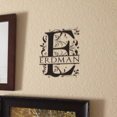 Adesivo decorativo preto com letra E e texto ERDMAN numa parede bege acima de fotografia em moldura preta.