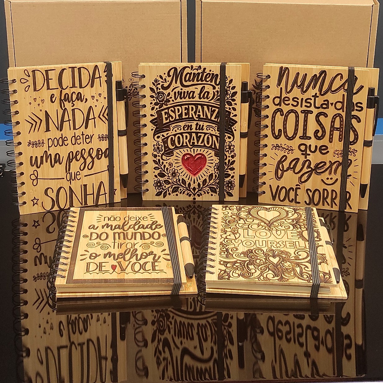 CADERNO ESPIRAL COM CAPA DE BAMBU E CANETA Cadernos de madeira com frases inspiradoras e canetas, dispostos em fundo preto refletivo.