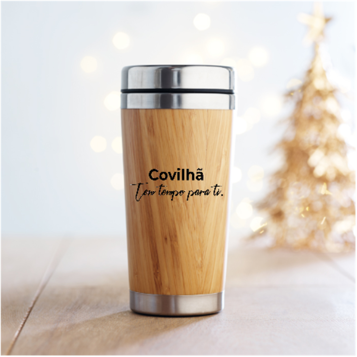 COPO DE AÇO INOX E BAMBÚ 500 ml Copo térmico de madeira com tampa metálica e texto impresso em ambiente com luzes e árvore dourada