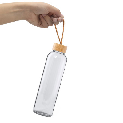 Garrafa de vidro transparente com tampa de madeira clara e cordão castanho, segurada por mão sobre fundo branco