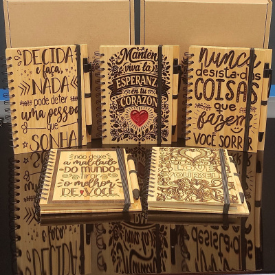 Cadernos de madeira com frases inspiradoras e canetas, dispostos em fundo preto refletivo.