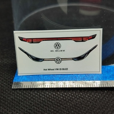 Etiqueta autocolante com ilustrações de luzes VW ID. BUZZ e texto sobre superfície preta e azul junto a régua.
