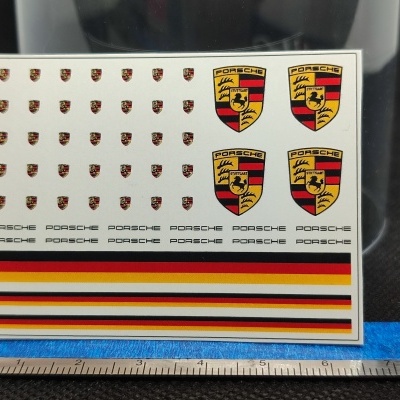 Folha de autocolantes com logótipos Porsche e faixas coloridas colocada sobre superfície preta com régua.