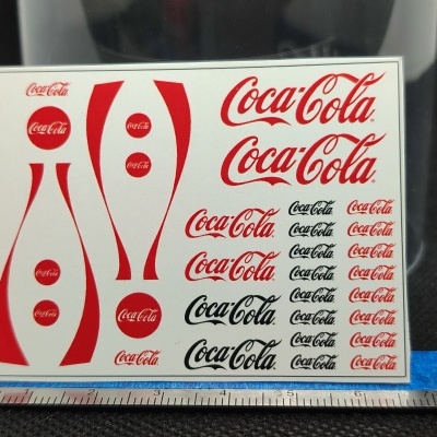 Folha com decalques Coca Cola vermelhos e pretos em superfície preta com régua metálica.