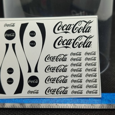 Folha de decalques Coca-Cola preta sobre fundo branco com régua e superfície azul em fundo