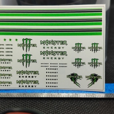 Folha branca com autocolantes verdes, pretos e brancos da marca MONSTER ENERGY com régua metálica abaixo.