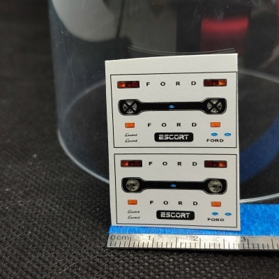 Folhas pequenas com autocolantes do Ford Escort colocadas sobre superfície preta com régua