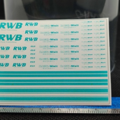 Folha com autocolantes RWB e RAUH-Welt BEGRIFF em azul e branco sobre régua em centímetros.