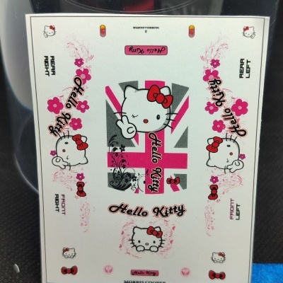 Folha de autocolantes Hello Kitty com flores rosa, laços vermelhos e texto preto ao lado de uma régua metálica na superfície preta.