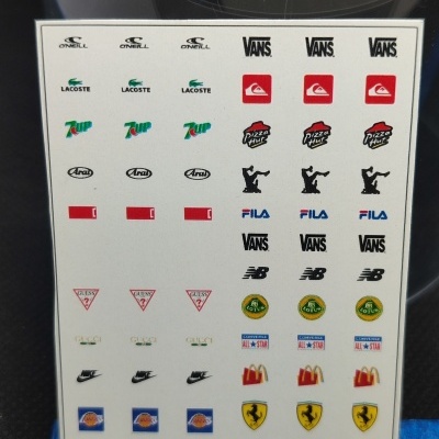 Folha branca com autocolantes pequenos de logos de marcas famosas e régua em fundo escuro.