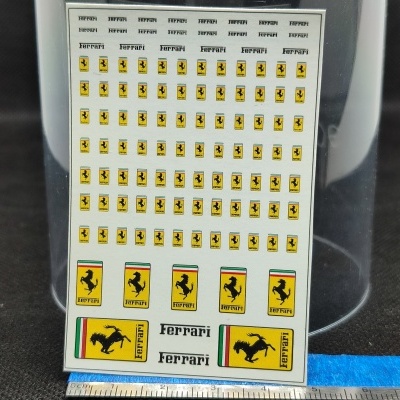 Folha branca com autocolantes do logótipo da Ferrari em várias dimensões, colocada sobre superfície escura com régua em baixo.
