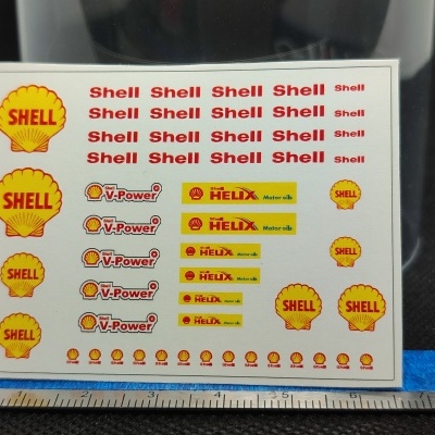 Folha de autocolantes com logotipos da Shell em cores amarelo, vermelho, preto e branco, sobre fundo preto e régua azul em baixo.