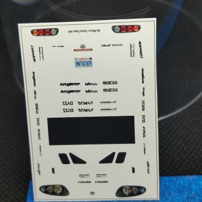 Folha de autocolantes para modelo de carro com detalhes e logotipos variados sobre superfície preta e azul com régua em centímetros