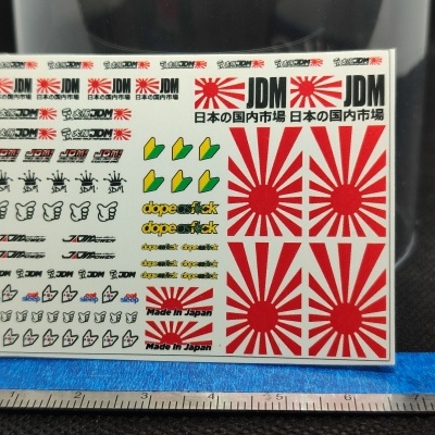 Folha de autocolantes JDM coloridos em papel com diversos símbolos, textos e ícones, com régua abaixo.