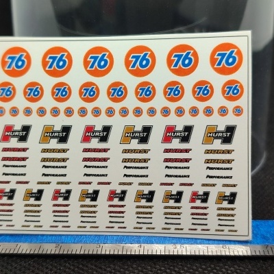Folha de autocolantes com logos 76 e HURST em fundo branco sobre superfície preta com régua