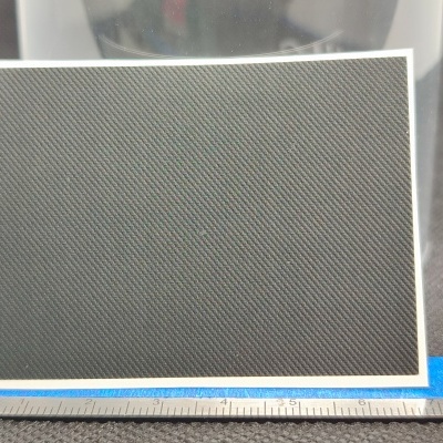 Painel retangular cinzento escuro com padrão diagonal e moldura branca sobre fita azul com régua em centímetros.