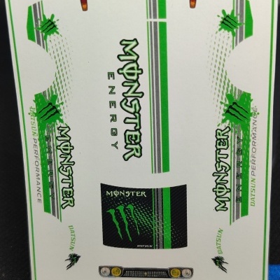 Folha de autocolantes brancos com logos Monster Energy e DATSUN PERFORMANCE em verde e preto ao lado de régua
