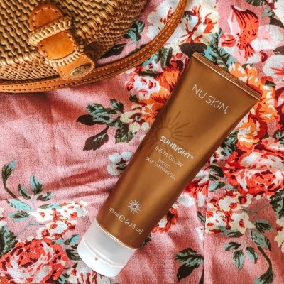 Tubo de gel autobronzeador NU SKIN SUNRIGHT INSTAGLOW sobre tecido floral rosa junto a mala de palha redonda.