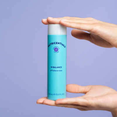 Frasco cilíndrico azul claro de toner entre mãos sobre fundo lilás