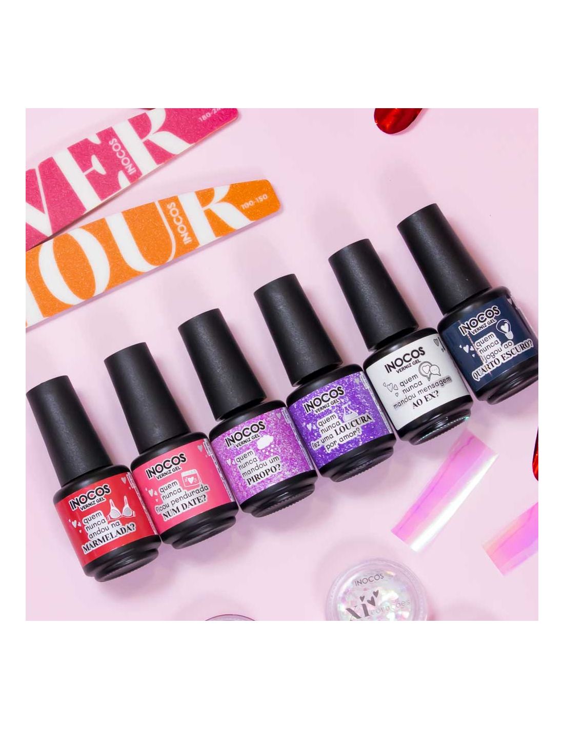 Coleção Vernizes de Gel Quem Nunca 15ml - Inocos Seis frascos de verniz de gel Nocos com cores variadas sobre mesa rosa com acessórios para unhas