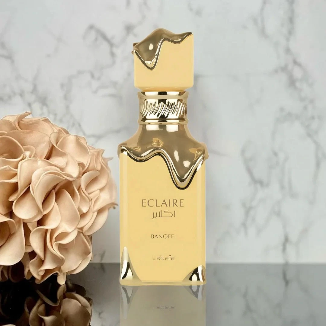 Eclaire Banoffi Lattafa Perfumes Compartilhável 100ml frasco amarelo de perfume dourado com flor decorativa ao lado em bancada preta refletora