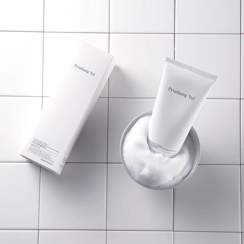 Pyunkang Yul Cleansing Foam Produto de limpeza facial Pyunkang Yul com embalagem branca e espuma em azulejos brancos
