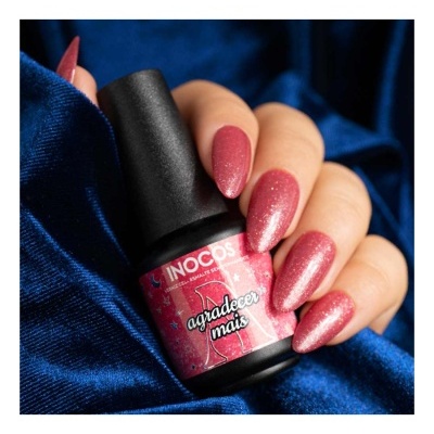 Frasco de verniz de unhas INOCOS rosa com glitter nas mãos, fundo de tecido azul